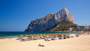 Een strand met ligbedden en parasols, een grote rotsformatie op de achtergrond en een helderblauwe lucht.