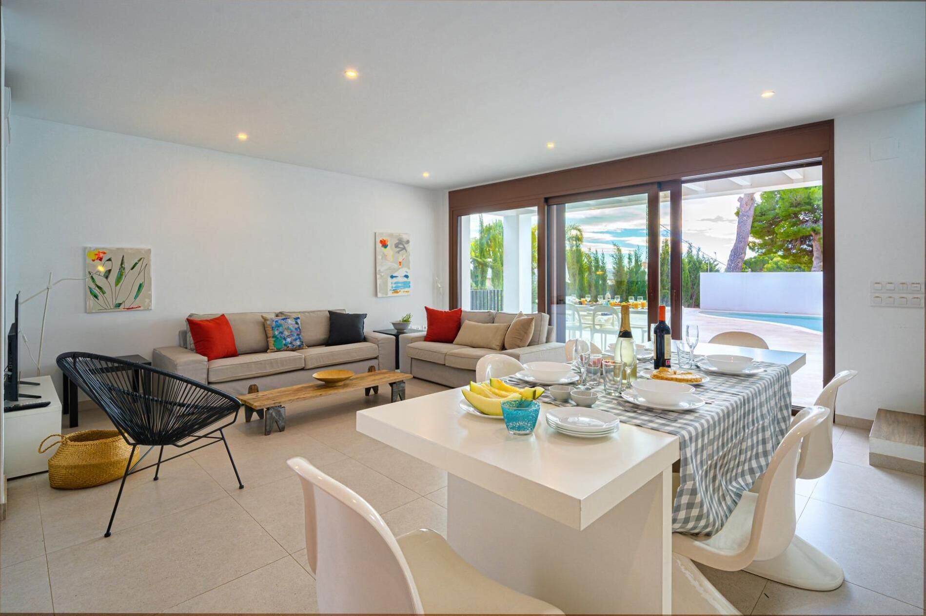 Ein modernes Wohnzimmer mit einer Couch, einem Esstisch und Stühlen. Ein großes Fenster bietet Blick auf einen Pool und viel Grün.