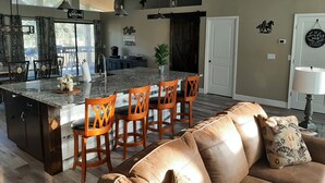 A spacious living area with a large brown leather sofa, a kitchen with a central island, wooden chairs, and pendant lighting.