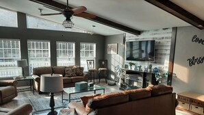 A modern living room with a brown leather sofa, a glass coffee table, and a television mounted on the wall. There are multiple windows with blinds, a ceiling fan, and a fireplace.
