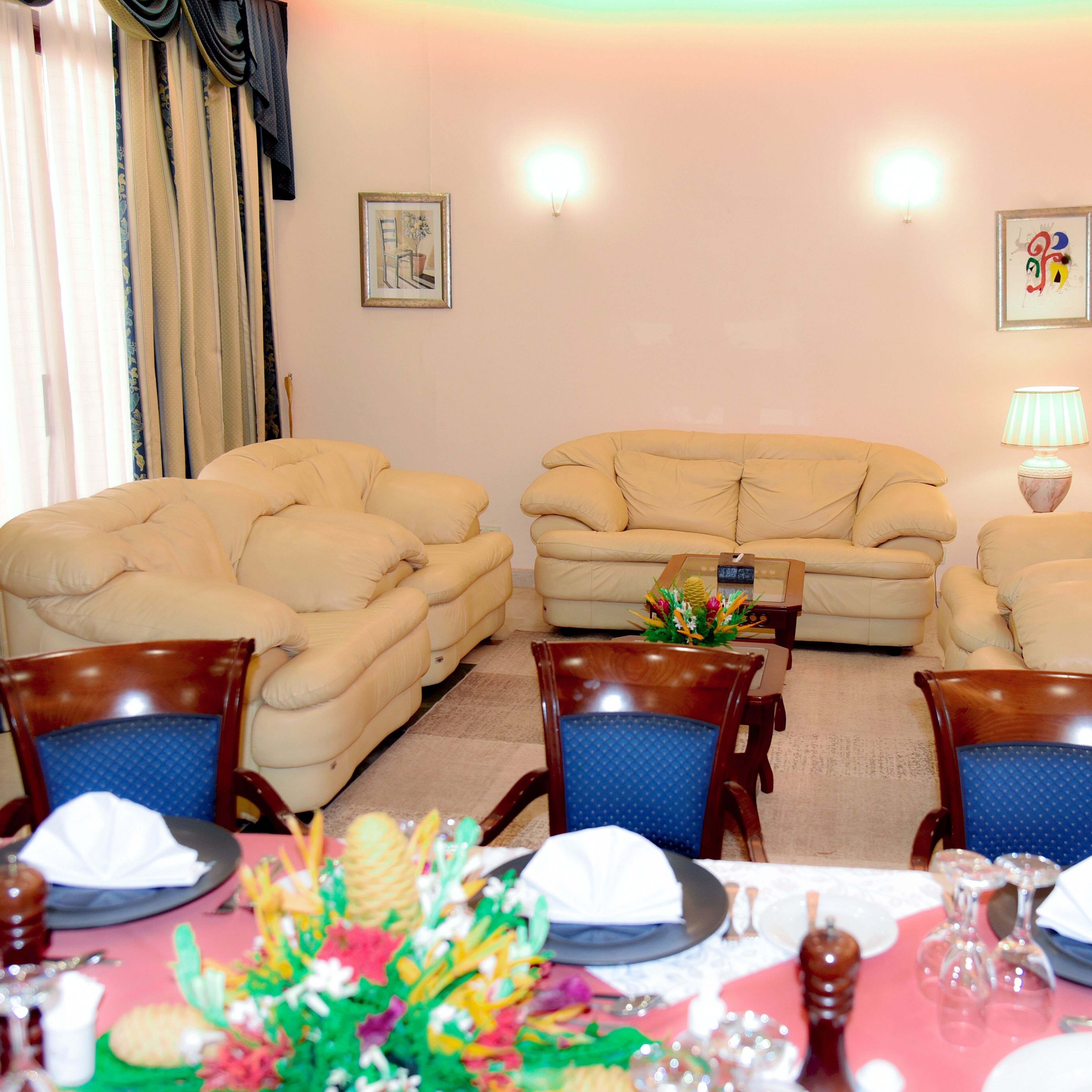 A living room with a beige sofa set, wooden chairs, a dining table set for four, and a small coffee table with a floral centerpiece.