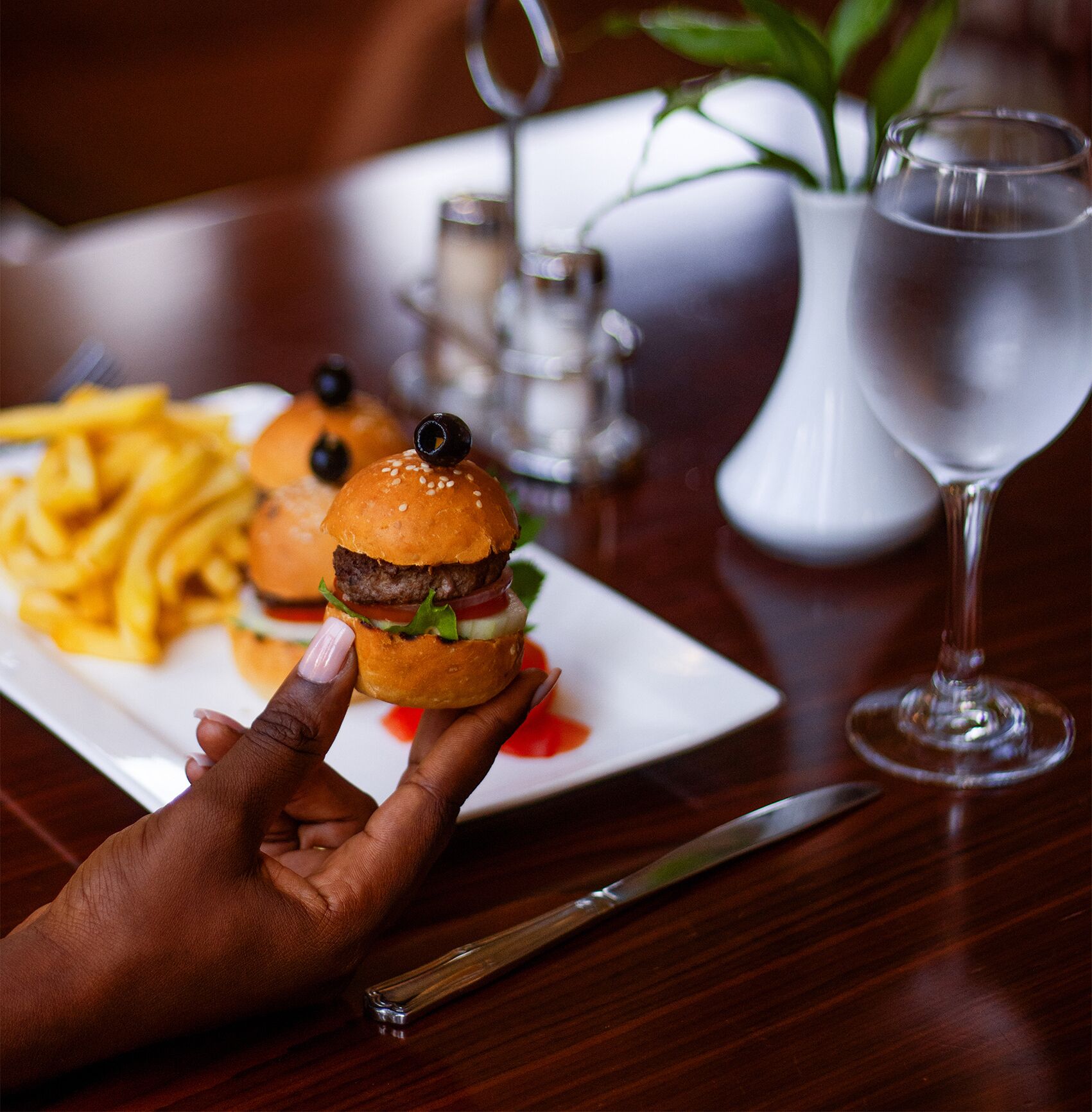 A hand holding a burger with a bun, lettuce, tomato, and a sesame seed bun, next to a plate of fries and a glass of water on a wooden table.