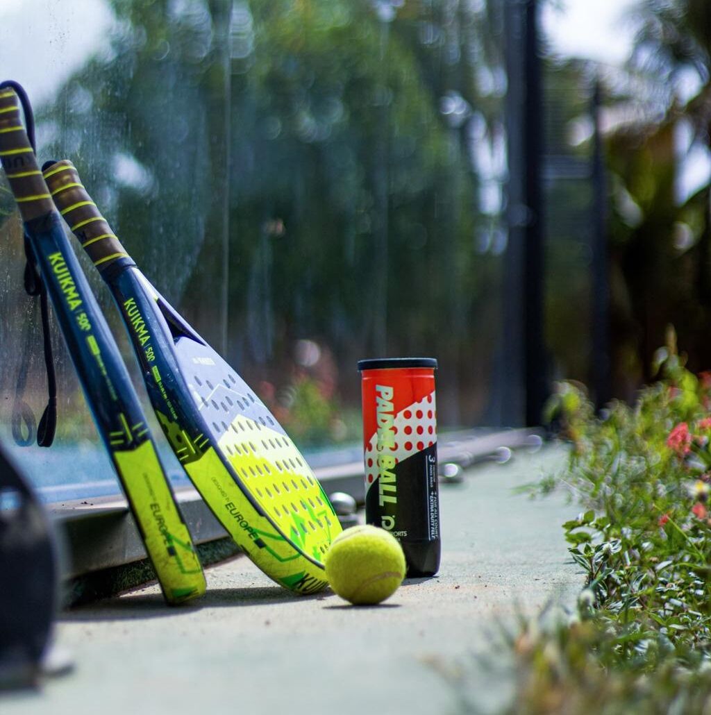 A yellow tennis ball, two tennis rackets, and a can of Pabst Blue Ribbon beer on a concrete surface.