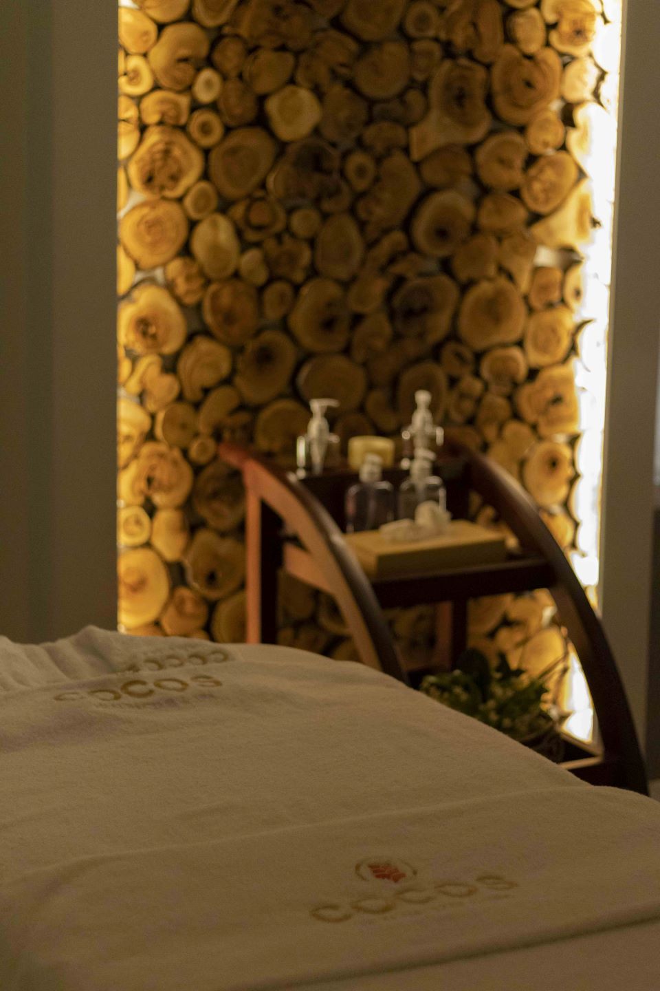A bed with a white cover and a visible logo, a wooden chair, and a wall decorated with wooden logs.