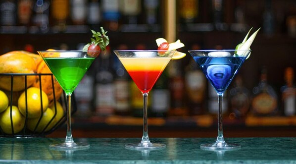 Three colorful cocktails in martini glasses with garnishes, placed on a bar counter with a blurred background of bottles. - Lancaster Ouaga 2000 (Ouagadougou)