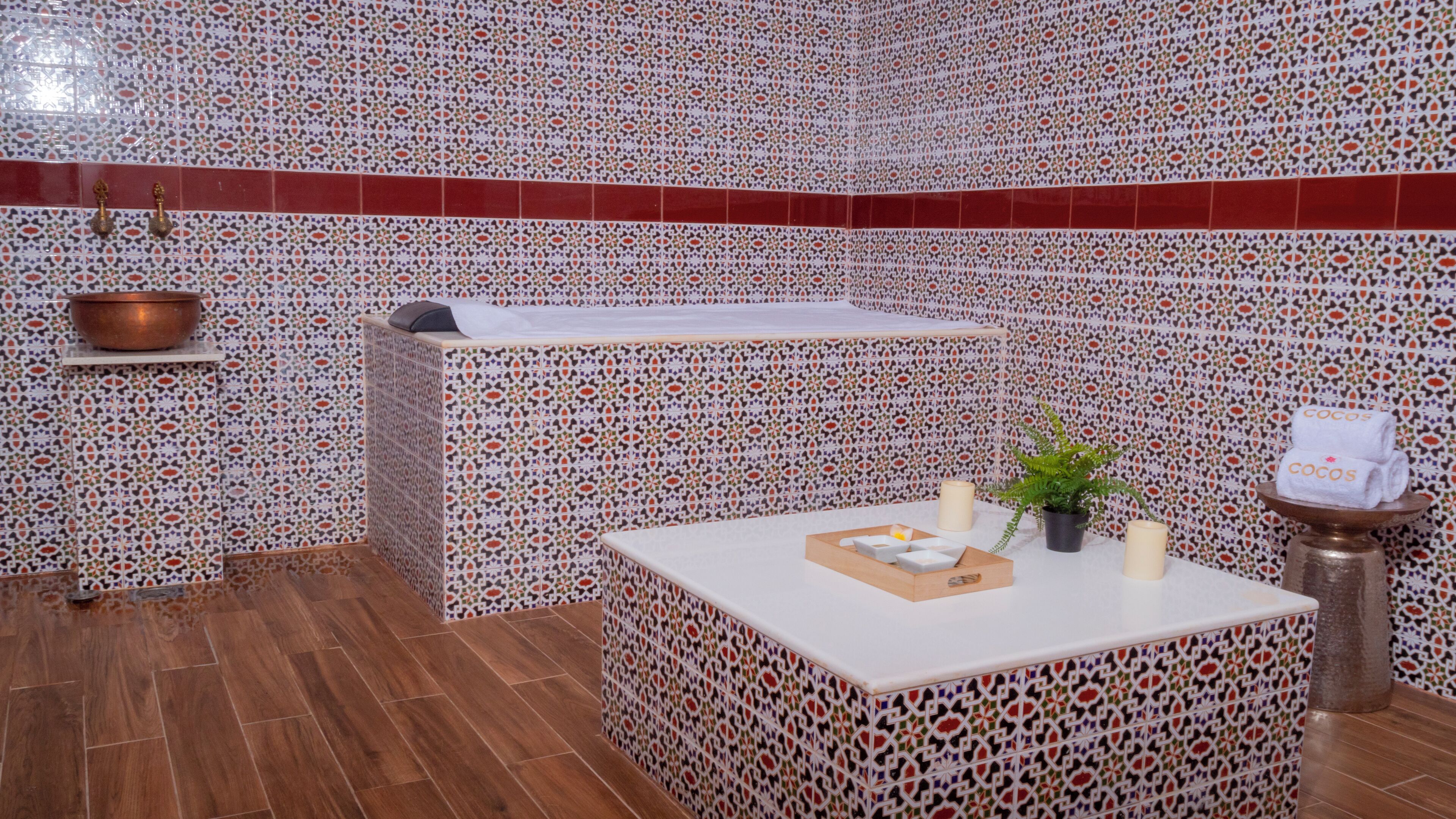 Casa de banho com parede de azulejos estampados, chão de madeira, banheira, lavatório, porta-toalhas e uma pequena planta.