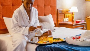 Una persona con bata blanca sentada en una cama con una bandeja de desayuno que incluye huevos, tostadas y fruta.