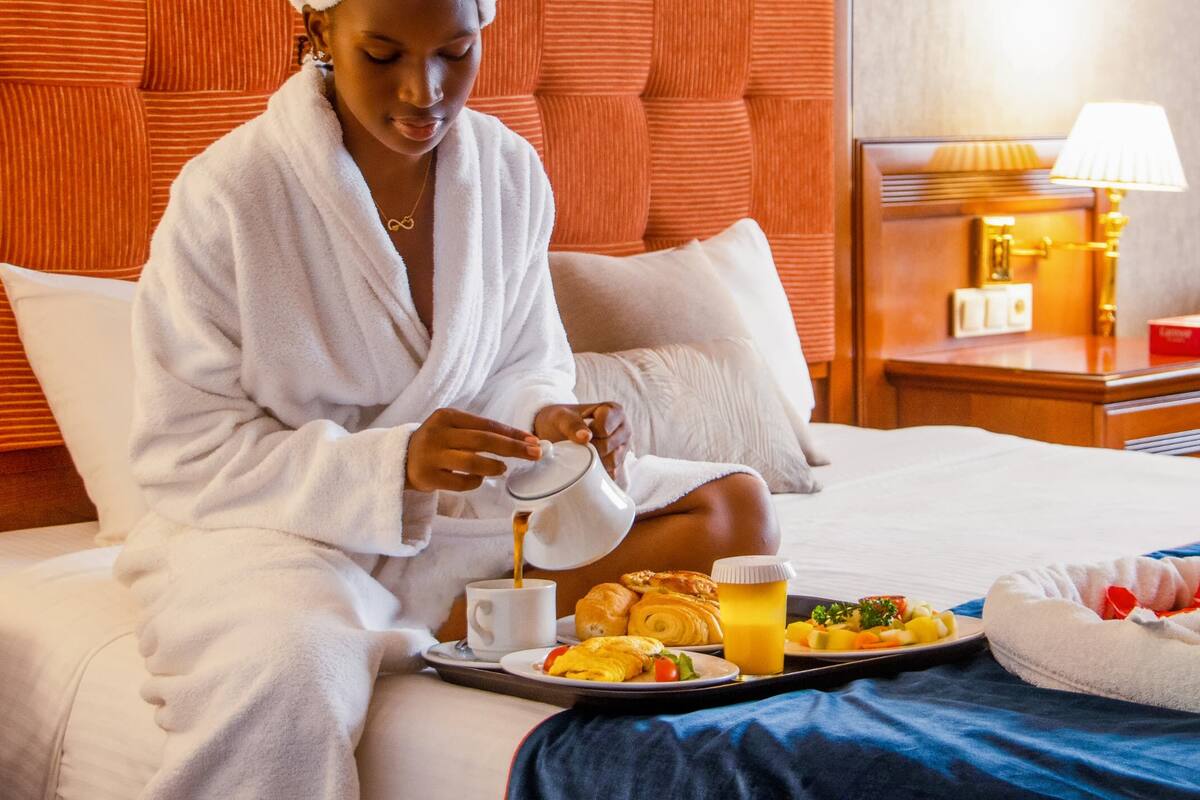 Eine Person in einem weißen Bademantel sitzt auf einem Bett mit einem Tablett mit Frühstücksartikeln, darunter Eier, Toast und Obst.