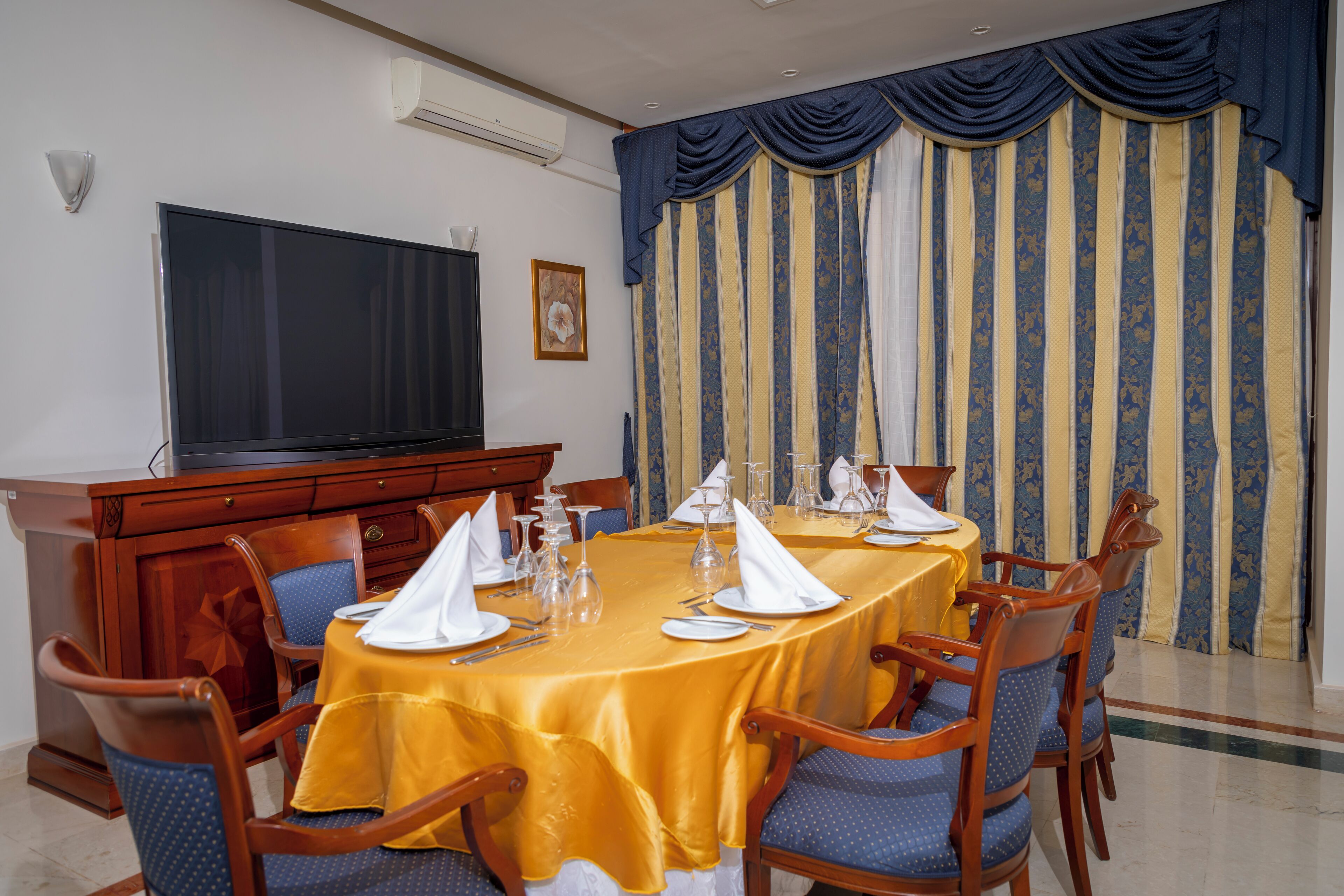 Un comedor con una mesa grande dispuesta para una comida, rodeada de sillas, y un televisor en la pared.