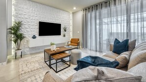 Un salon moderne doté d’un téléviseur à écran plat fixé sur un mur à texture, d’un confortable ensemble de canapés, d’une table basse et d’une grande fenêtre ornée de rideaux légers.
