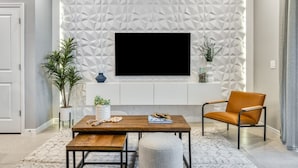 Un salon moderne avec un téléviseur à écran plat fixé à un mur orné d’un motif géométrique, une table basse en bois, un fauteuil en cuir et une plante en pot.