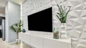 Un salon moderne avec un téléviseur à écran plat fixé sur une console blanche, un mur orné de motifs géométriques et des plantes décoratives.