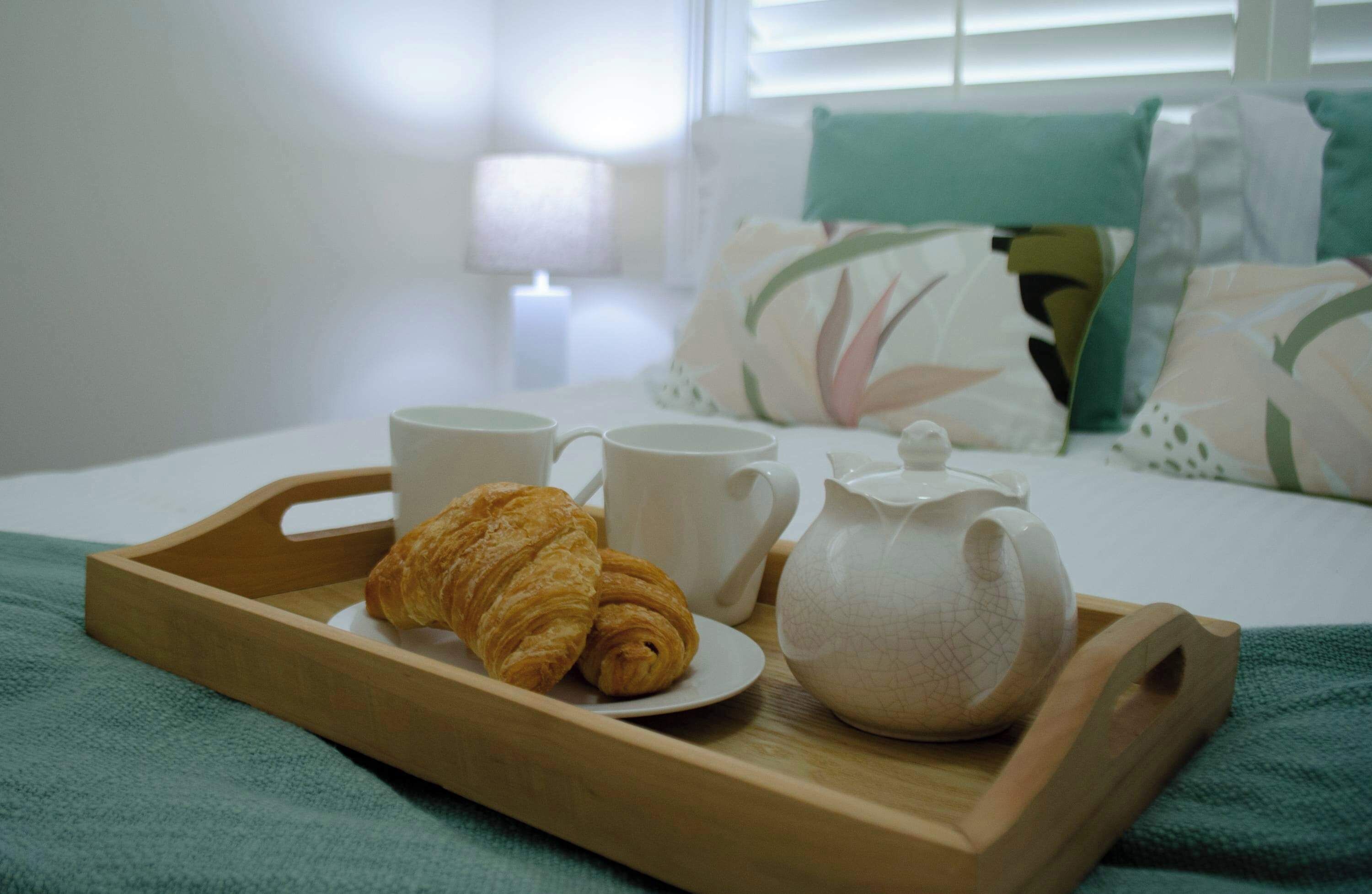 Un vassoio di legno con croissant, una teiera e due tazze su un letto con una coperta verde.