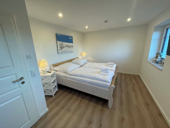A bedroom with a wooden bed, white bedding, two bedside tables with lamps, a wall-mounted painting, and a window with a view.
