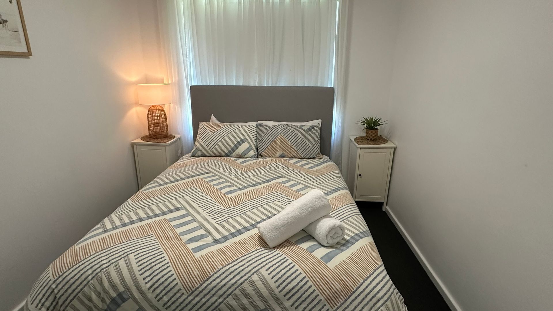Een net opgemaakt bed met behang, twee nachtkastjes met lampen, en een raam met dunne gordijnen.