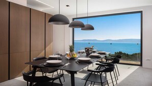A modern dining area with a large window overlooking the sea, a minimalist table setup, and pendant lighting. - Villa Senzea  - modern villa with heated pool, gym, sauna, jacuzzi and sea views by MyWaycation (Marusici)