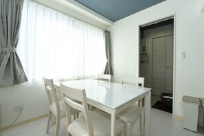 Una habitación con una mesa y sillas blancas, una ventana con cortinas, una puerta y un pequeño armario blanco.