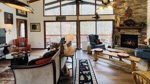 Ein geräumiges Wohnzimmer mit hohen Decken, Holzbalken, einem Kamin aus Stein und großen Fenstern mit Blick ins Freie.