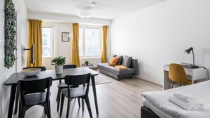 Moderni, hyvin valaistu huone, jossa on ruokapöytä, tuoleja, sohva, sänky ja työpöytä tuolilla.