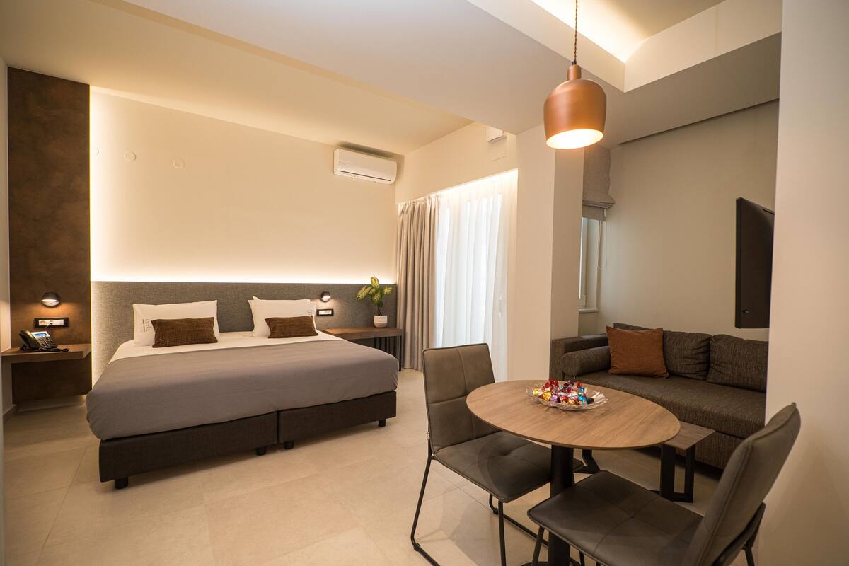 A modern hotel room with a large bed, a sofa, a dining table, and a hanging pendant light.