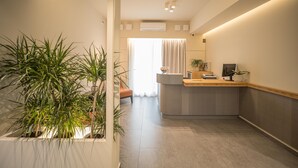 A modern reception area with a counter, potted plants, and a view of a room with a window and curtains.
