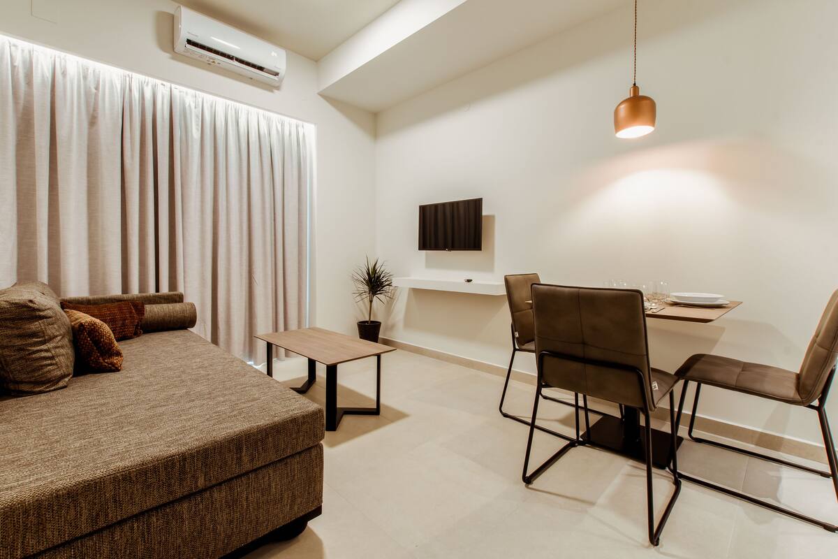 A modern living room with a sofa, a dining table with chairs, a TV mounted on the wall, and a hanging pendant light.