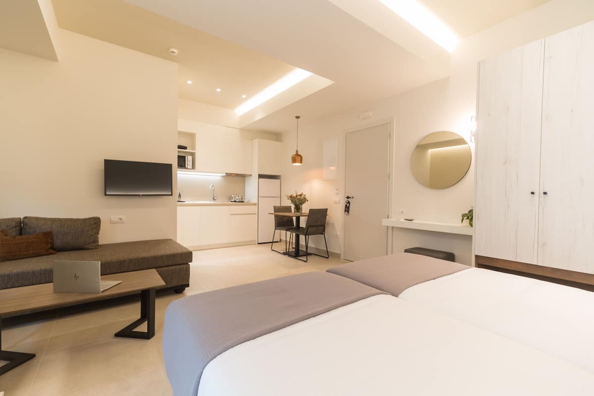 A modern hotel room with a large bed, a desk with a laptop, a sofa, and a kitchen area in the background.