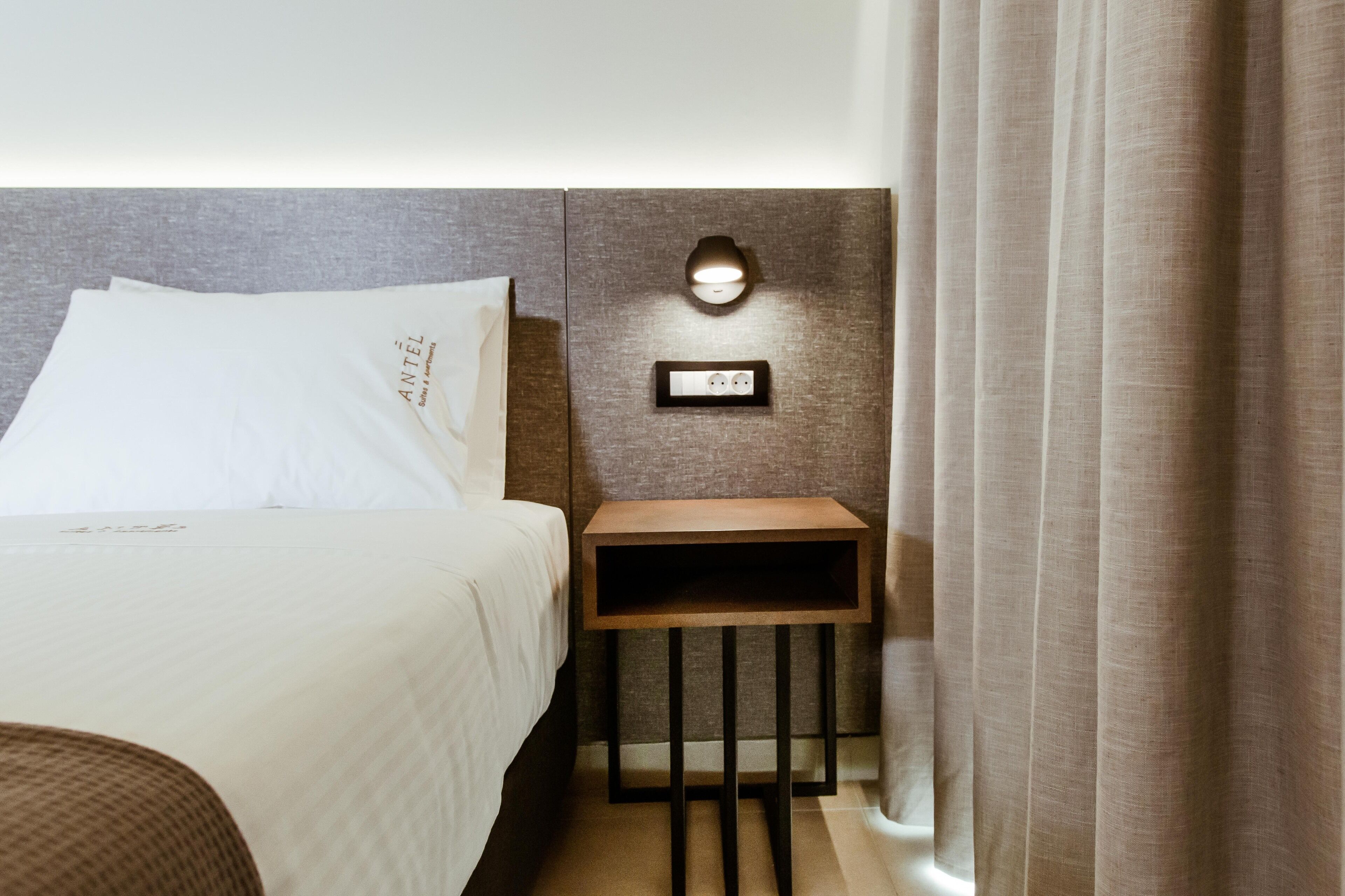A neatly made bed with white linens and a pillow with text, a small wooden nightstand with a wall-mounted light, and a power outlet.