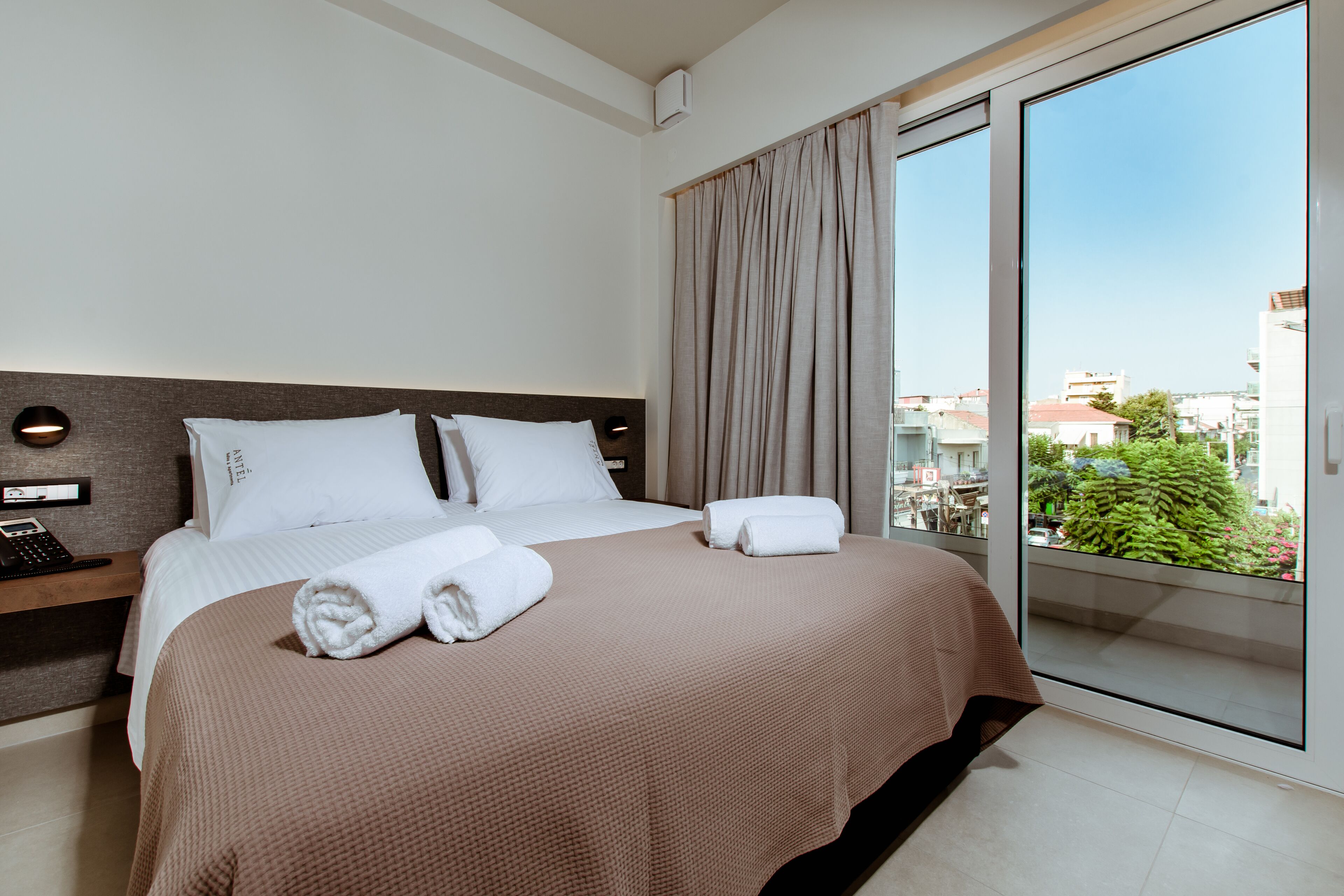 A hotel room with a large bed, a bedside table with a phone, and a view of the city through a large window.