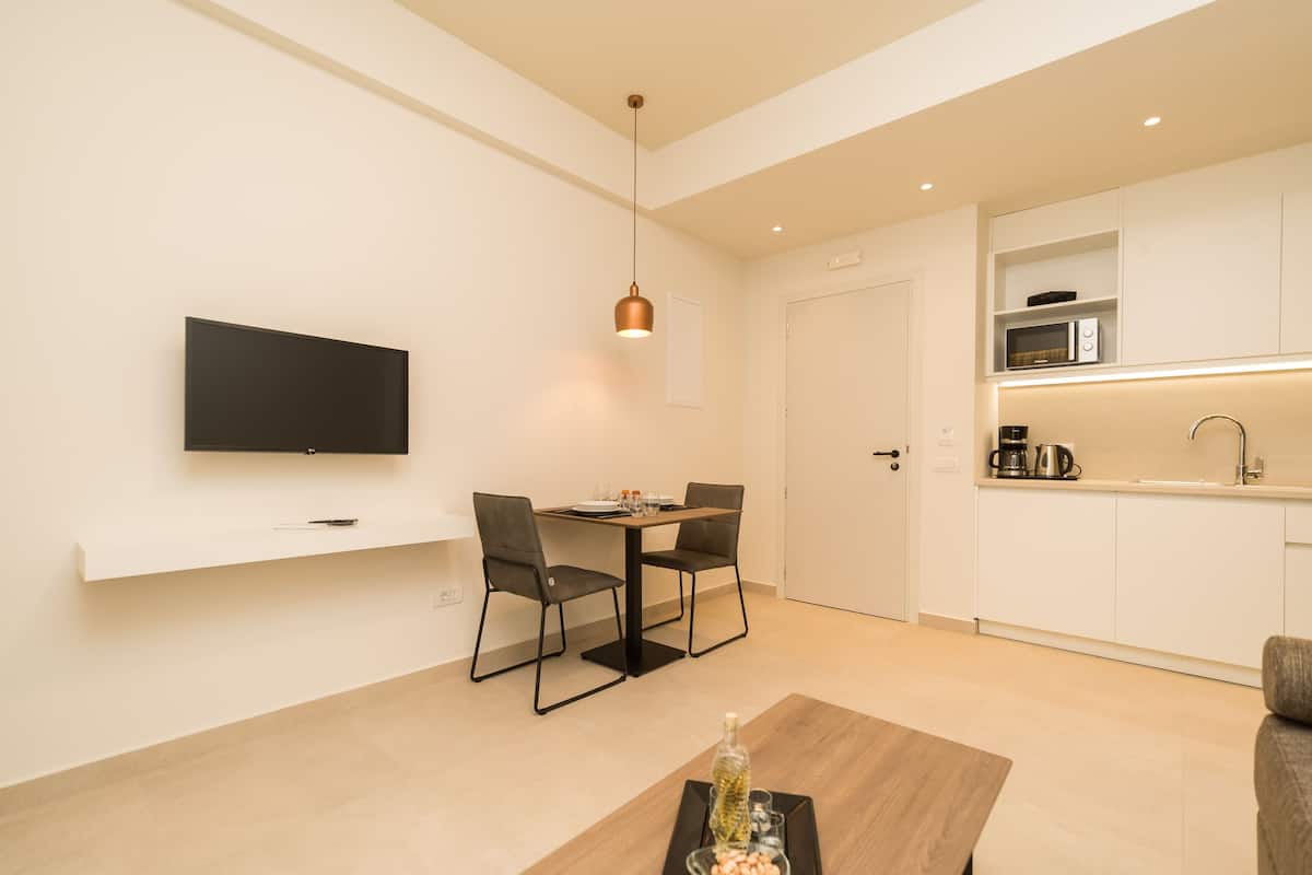 A modern living room with a flat-screen TV, a dining table with chairs, a coffee table, and a kitchen area with a microwave and coffee maker.