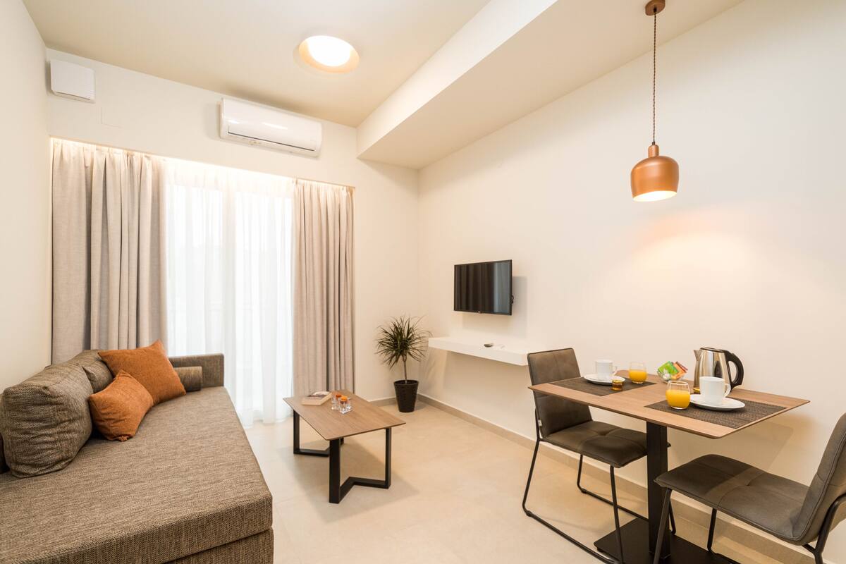 A modern living room with a sofa, a coffee table, a dining table with chairs, a TV mounted on the wall, and a potted plant.