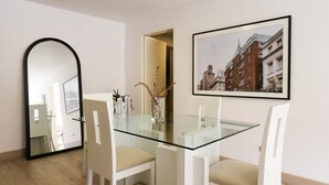 Un espace repas moderne avec une table en verre et des chaises blanches, un tableau représentant un paysage urbain encadré au mur, et un miroir qui reflète une partie de la pièce.