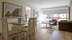Un salon moderne avec une table à manger en verre, deux canapés et une grande fenêtre équipée de stores.