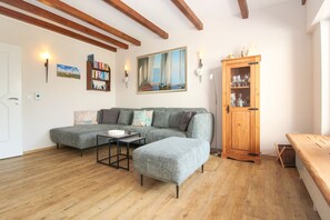 A living room with a sectional sofa, a small table, a wooden cabinet with glass doors, and a painting on the wall. - Leuchtfeuer Vacation Home in Greetsiel (Greetsiel)