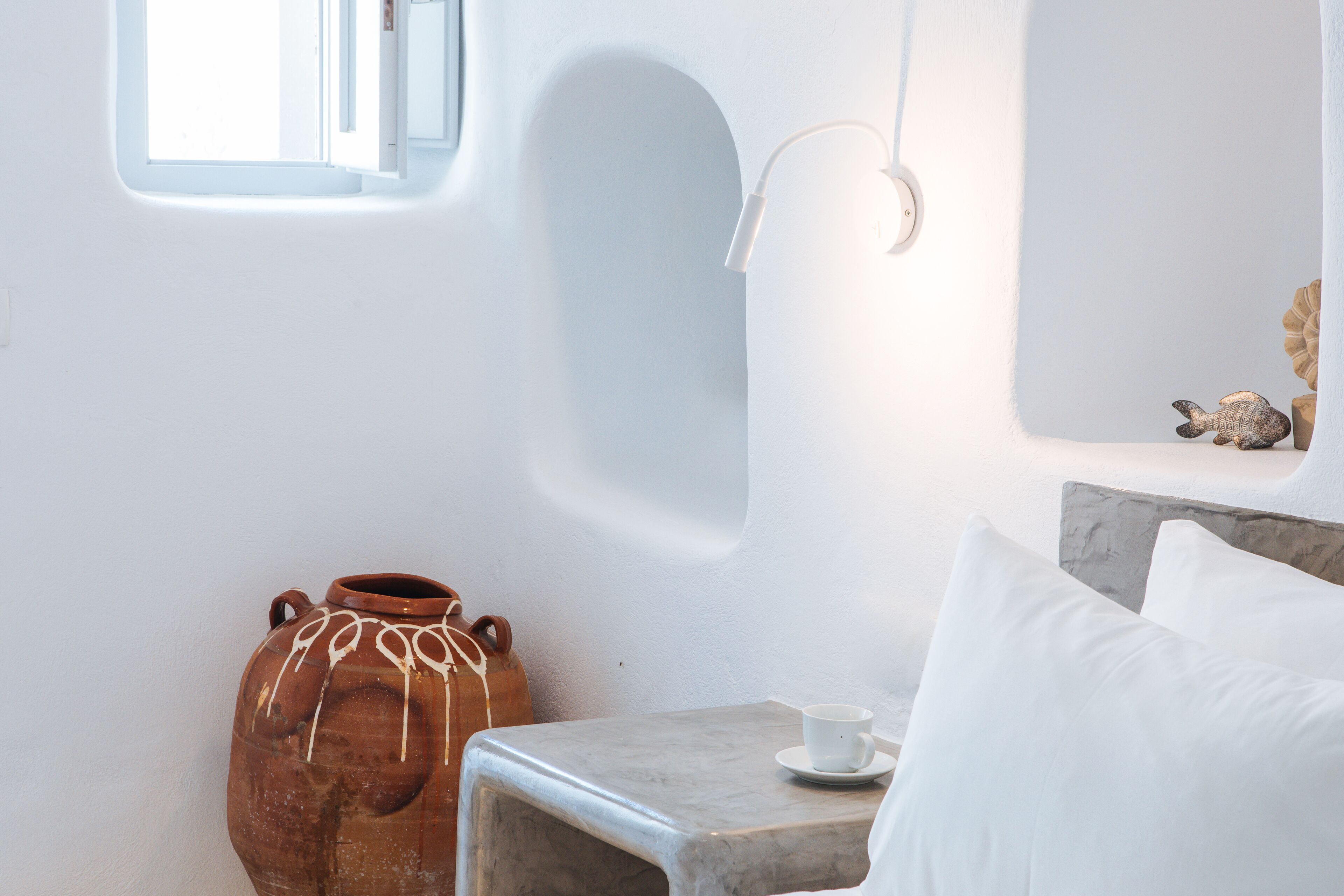 A room with a rustic clay pot, a small table with a cup, and a bed with white pillows.