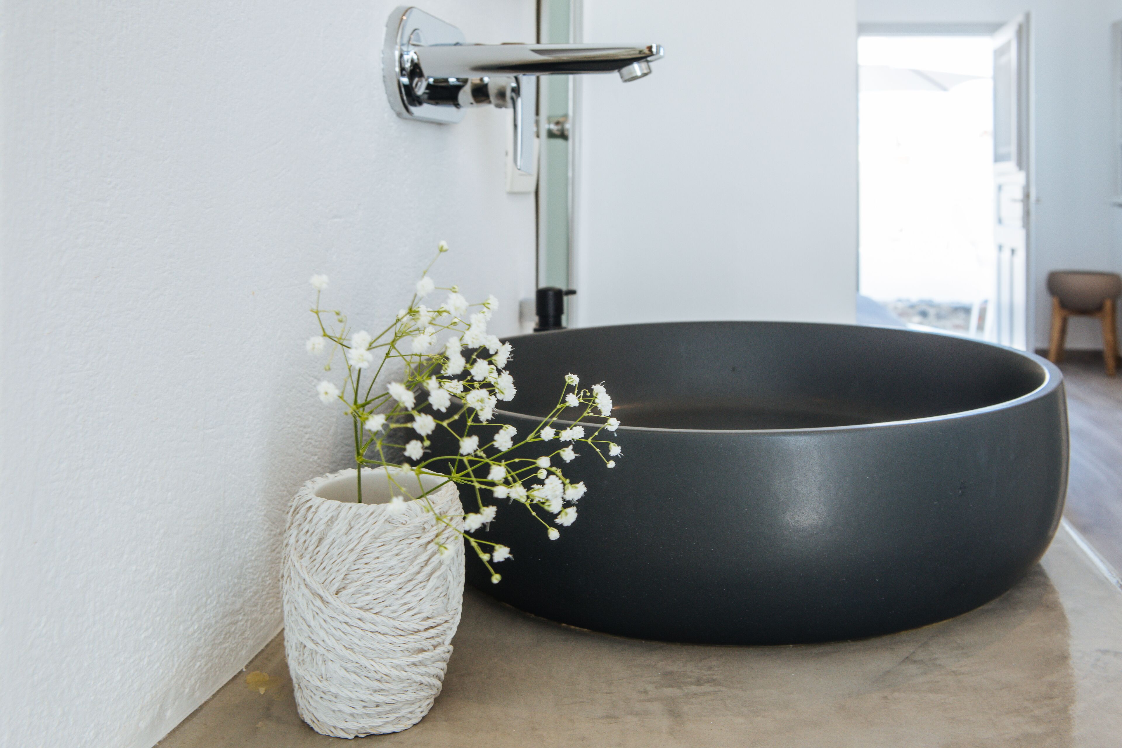 Ein Badezimmer mit einem runden, schwarzen Waschbecken, einer weißen gewobenen Vase mit Blumen und einem Chromarmaturenset.