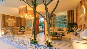 Un salon moderne avec un sol carrelé aux motifs géométriques, un ensemble de canapés et une table basse. Deux grands arbres se dressent au centre de la pièce, et l’ensemble dégage une atmosphère chaleureuse et accueillante grâce à un éclairage doux.