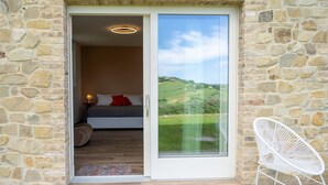 A bedroom with a stone wall, a bed with red pillows, a white chair, and a view of a green landscape. - Villa Edelia, Villa for exclusive use in Mondavio, Le Marche (Mondavio)