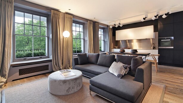 Uma sala de estar moderna com um sofá sectional cinza, um banquinho redondo e uma janela grande com cortinas.