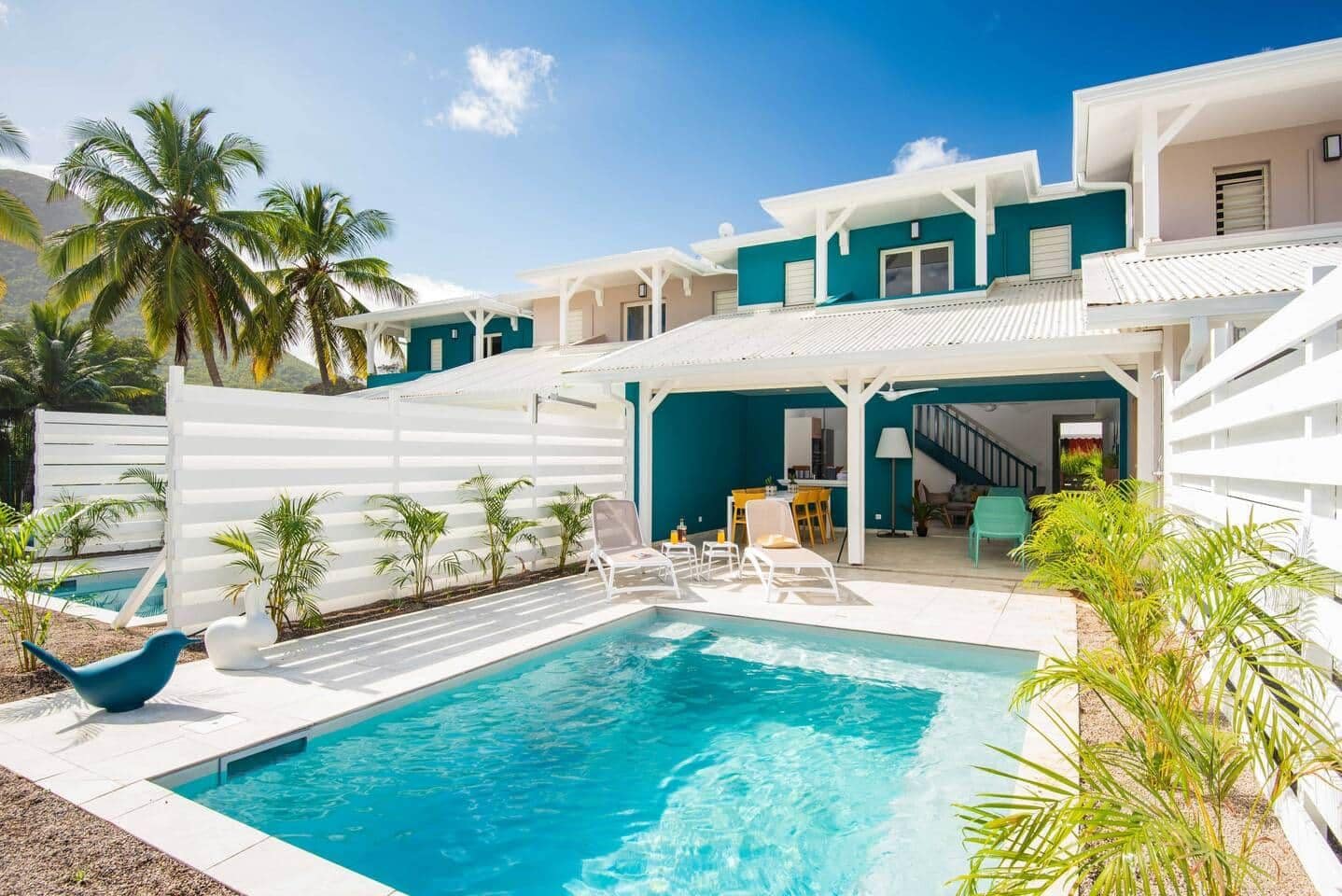 Un área de piscina moderna con una piscina azul cristalina, sillones blancos para tomar el sol y una zona de estar cubierta.