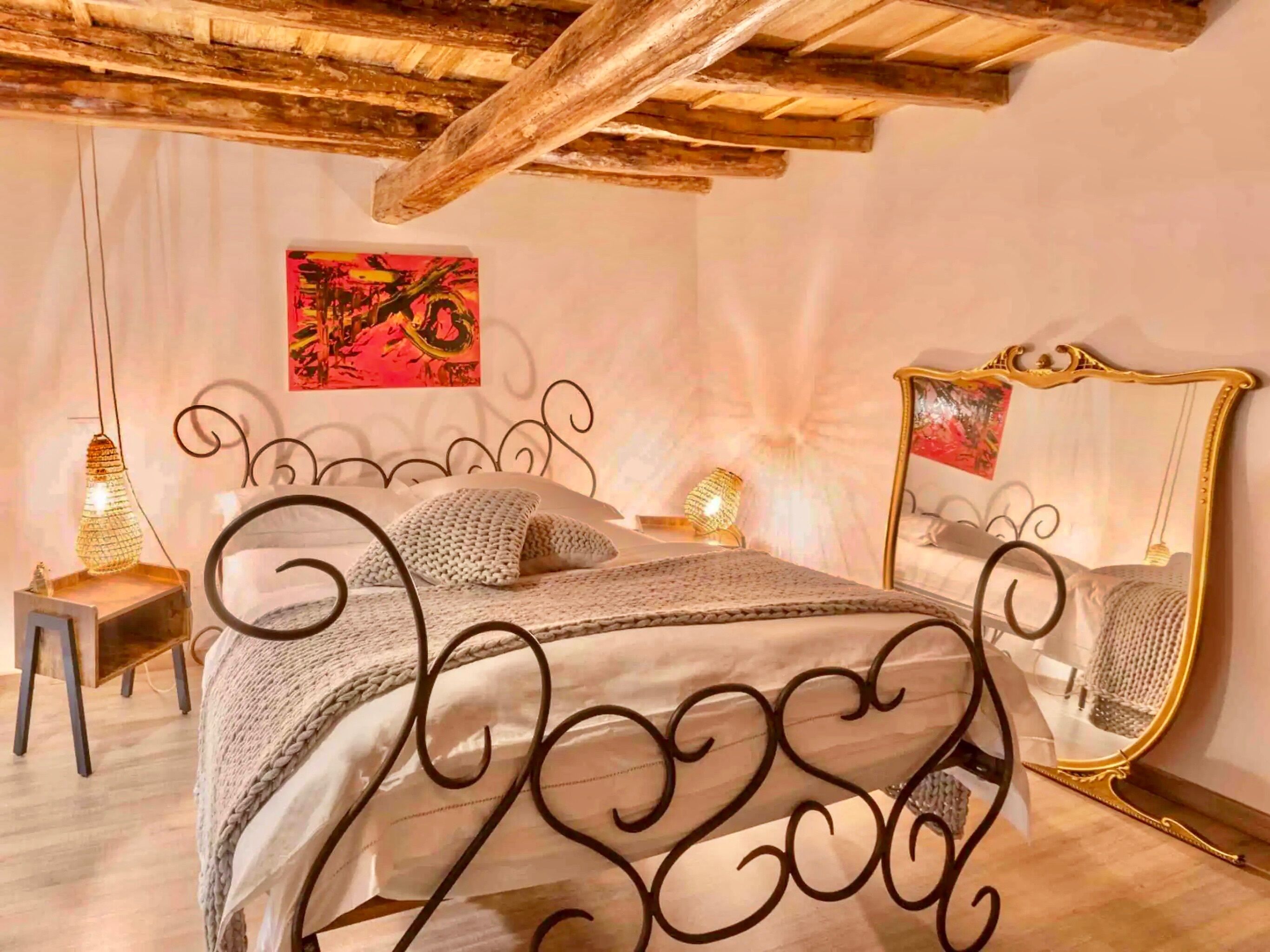 A bedroom with a metal bed frame, a wooden ceiling, a painting on the wall, and a mirror with a gold frame.