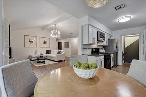 A modern kitchen with white cabinets, a dining table with a bowl of apples, and a living area with a sofa and coffee table. - Stylish 3BR | King Bed | ACL x Formula 1 (Austin)