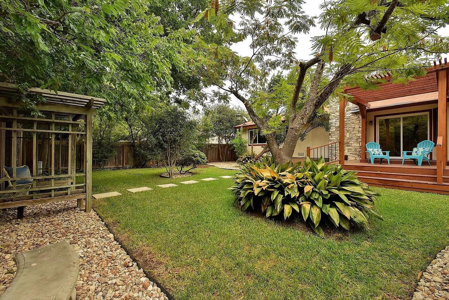 A well-maintained backyard with a wooden pergola, a seating area with chairs, and a stone building with a glass door.