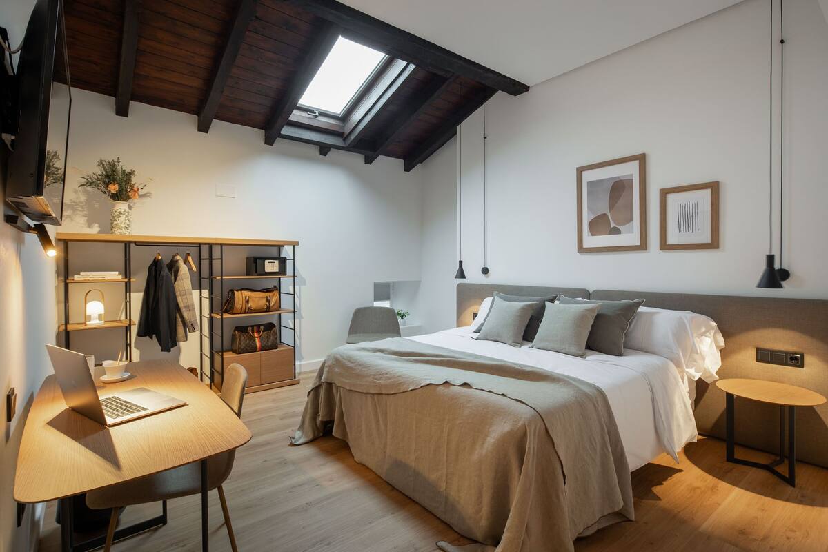 A modern bedroom with a skylight, a bed with a headboard, a desk with a laptop, and a wardrobe.