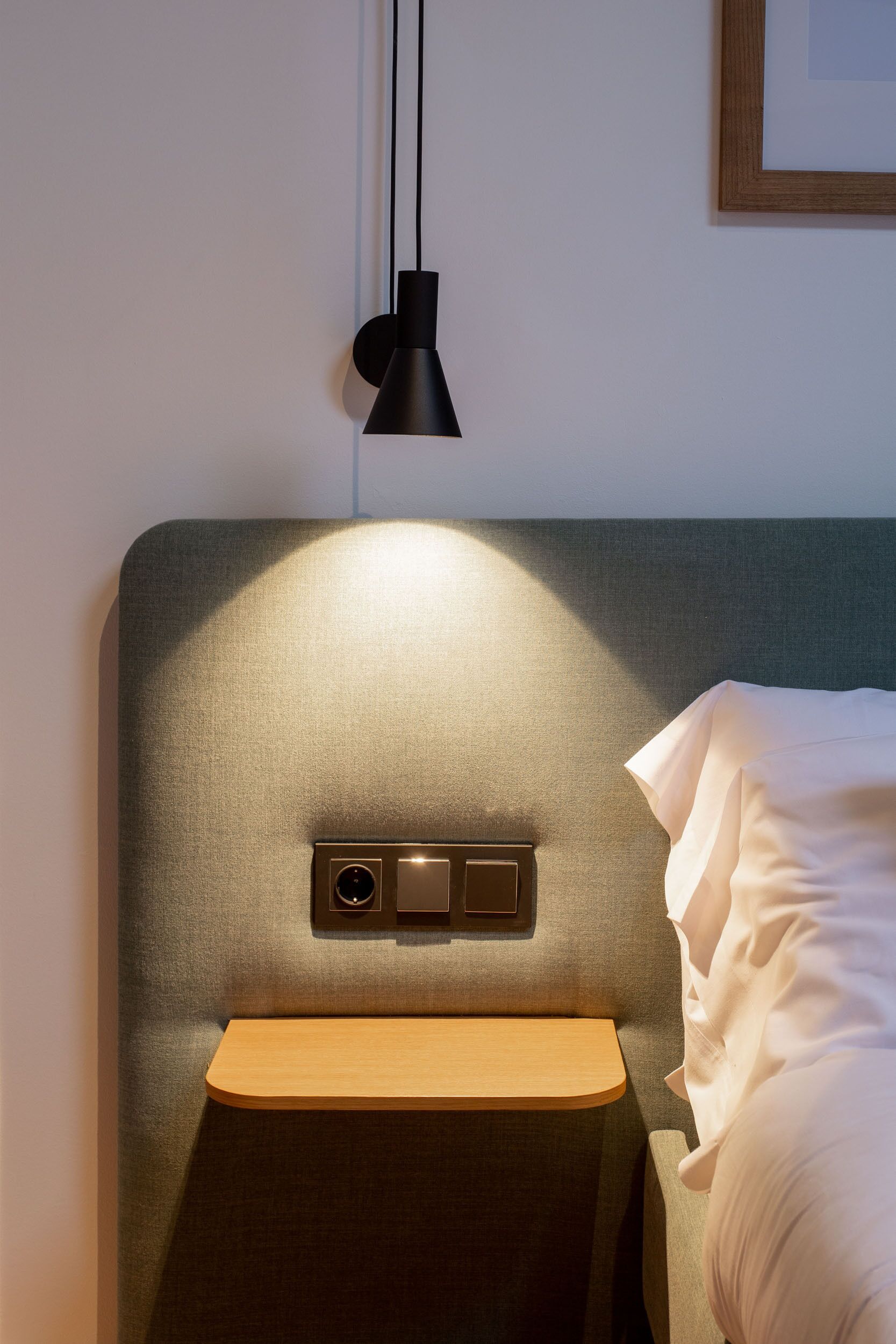 A neatly made bed with a wooden nightstand, a wall-mounted control panel, and a pendant light.