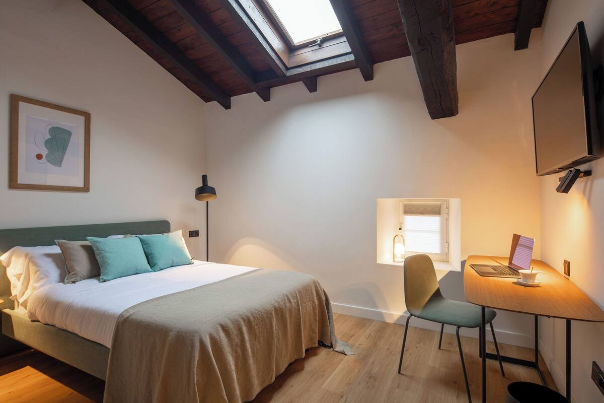 A bedroom with a bed, a desk, a chair, a window, and a painting on the wall.