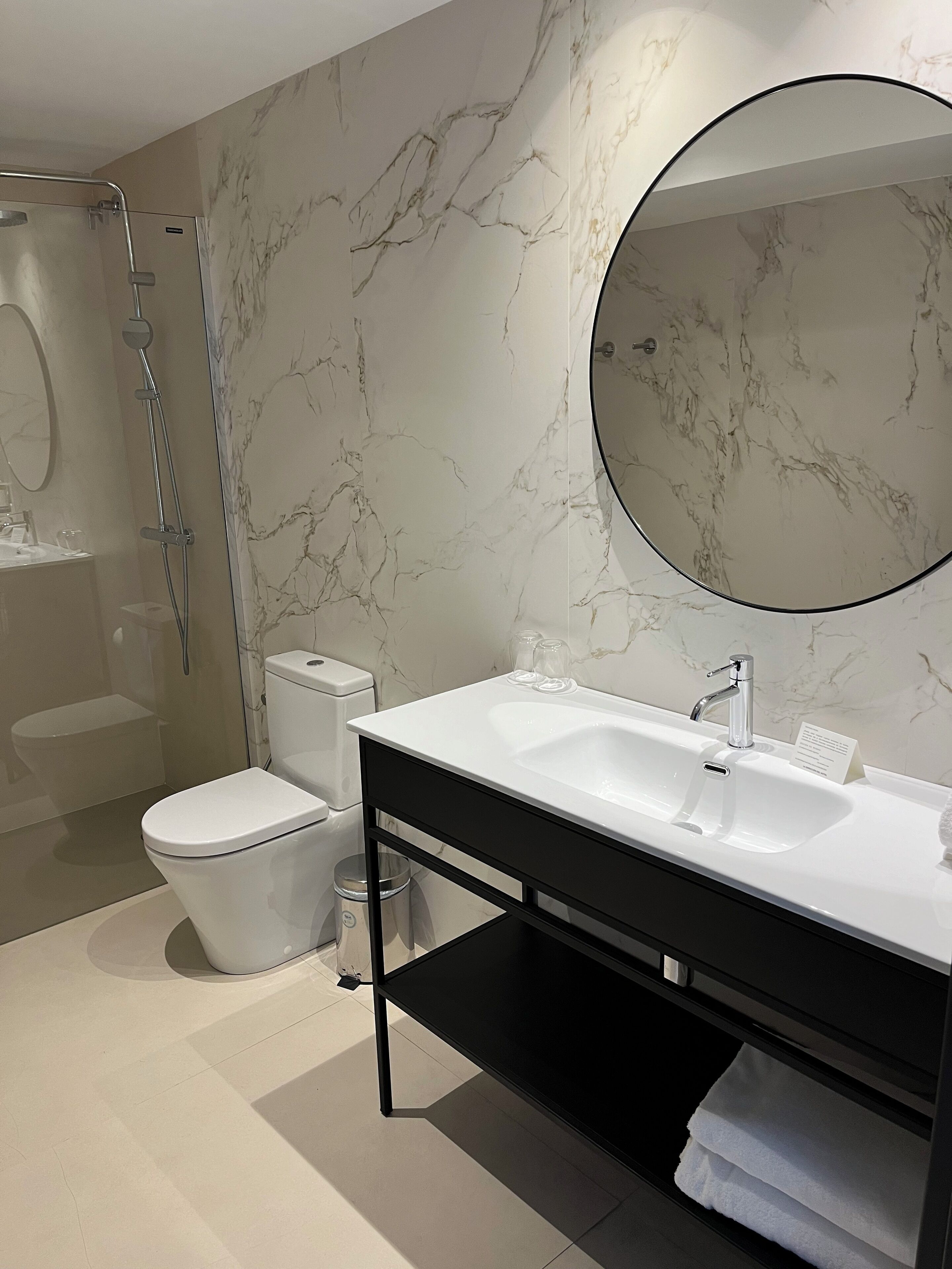 A modern bathroom with a marble backsplash, a round mirror, a white sink, and a glass-enclosed shower.