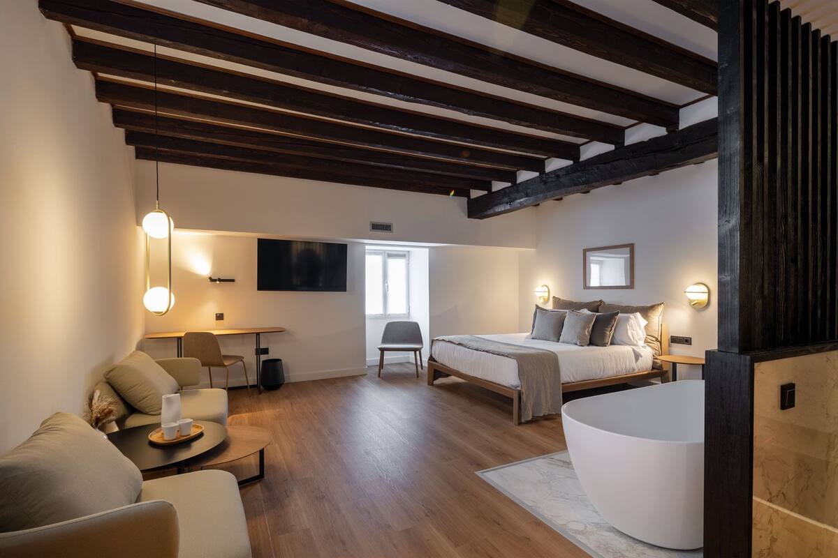 A modern hotel room with a wooden ceiling, a large bed, a seating area with a table, and a large window providing natural light.