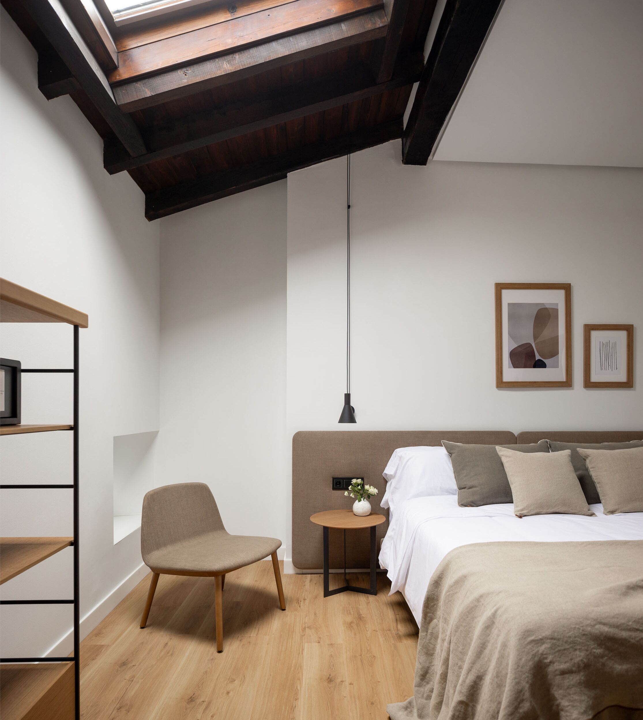 A bedroom with a skylight, a bed with pillows, a chair, a small table with a vase, and a shelf with books.