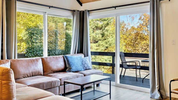 Een moderne woonkamer met een leren bank, een bijzettafeltje en grote ramen met uitzicht op een houten terras en bomen.